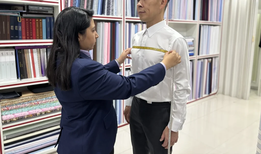 A tailor holding a measuring tape across a client’s chest in front of fabric shelves.