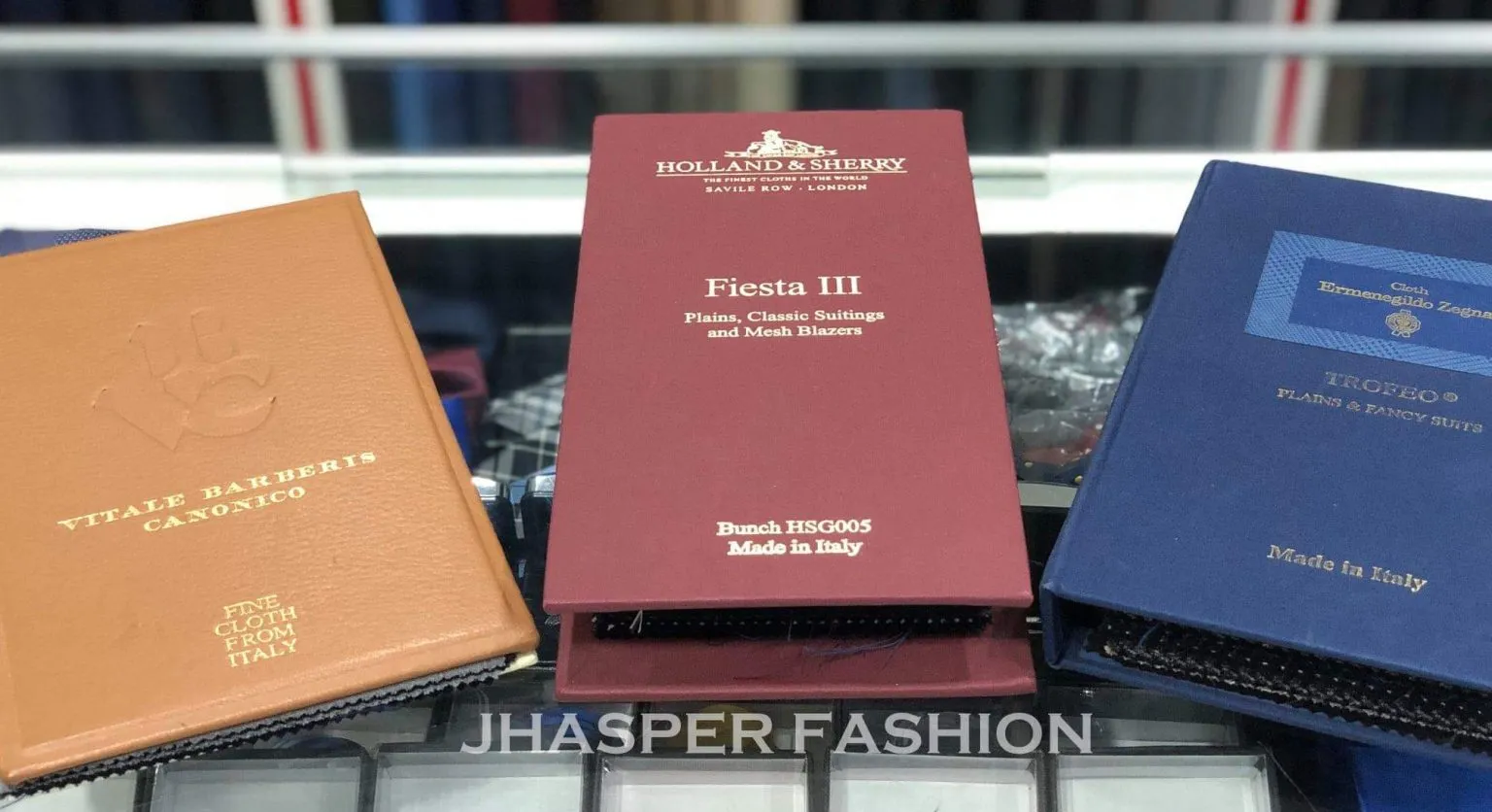 Suit fabric sample books displayed on a counter with Jhasper Fashion branding.