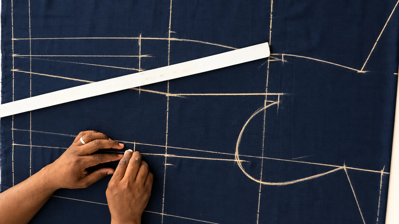 Hands marking garment pattern lines with chalk on navy fabric beside a ruler.